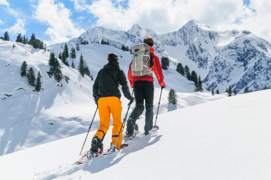 Zwei Menschen wandern mit Schneeschuhen an den Füßen über ein unverspurtes Schneefeld, im Hintergrund sind weitere Berge